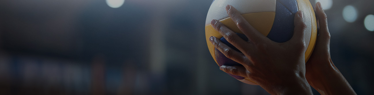 На изображении видны руки, держащие волейбольный мяч. Задний план размытый, светильники создают светлые круги на тёмном фоне